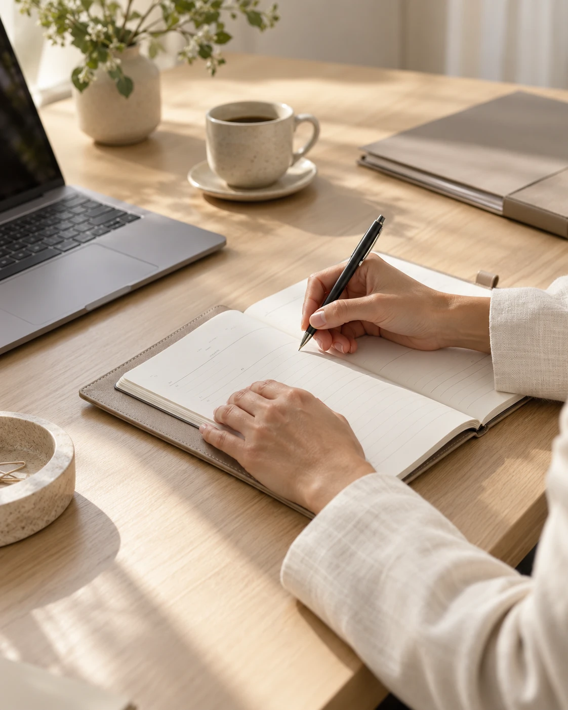 Hands writing in a planner at a sunlit desk, beside a laptop and a cup of coffee